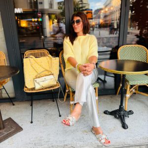 Frau in sommerlichem Outfit vor einem Straßencafé in Paris, trägt eine hellgelbe Bluse und silberne Hose. Neben ihr auf dem Stuhl liegt die große Boho Nomad – Citron in leuchtendem Gelb mit weißem Muster, handgefertigt in Marokko.
