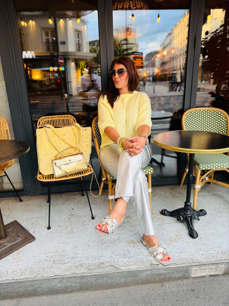 Frau in sommerlichem Outfit vor einem Straßencafé in Paris, trägt eine hellgelbe Bluse und silberne Hose. Neben ihr auf dem Stuhl liegt die große Boho Nomad – Citron in leuchtendem Gelb mit weißem Muster, handgefertigt in Marokko.
